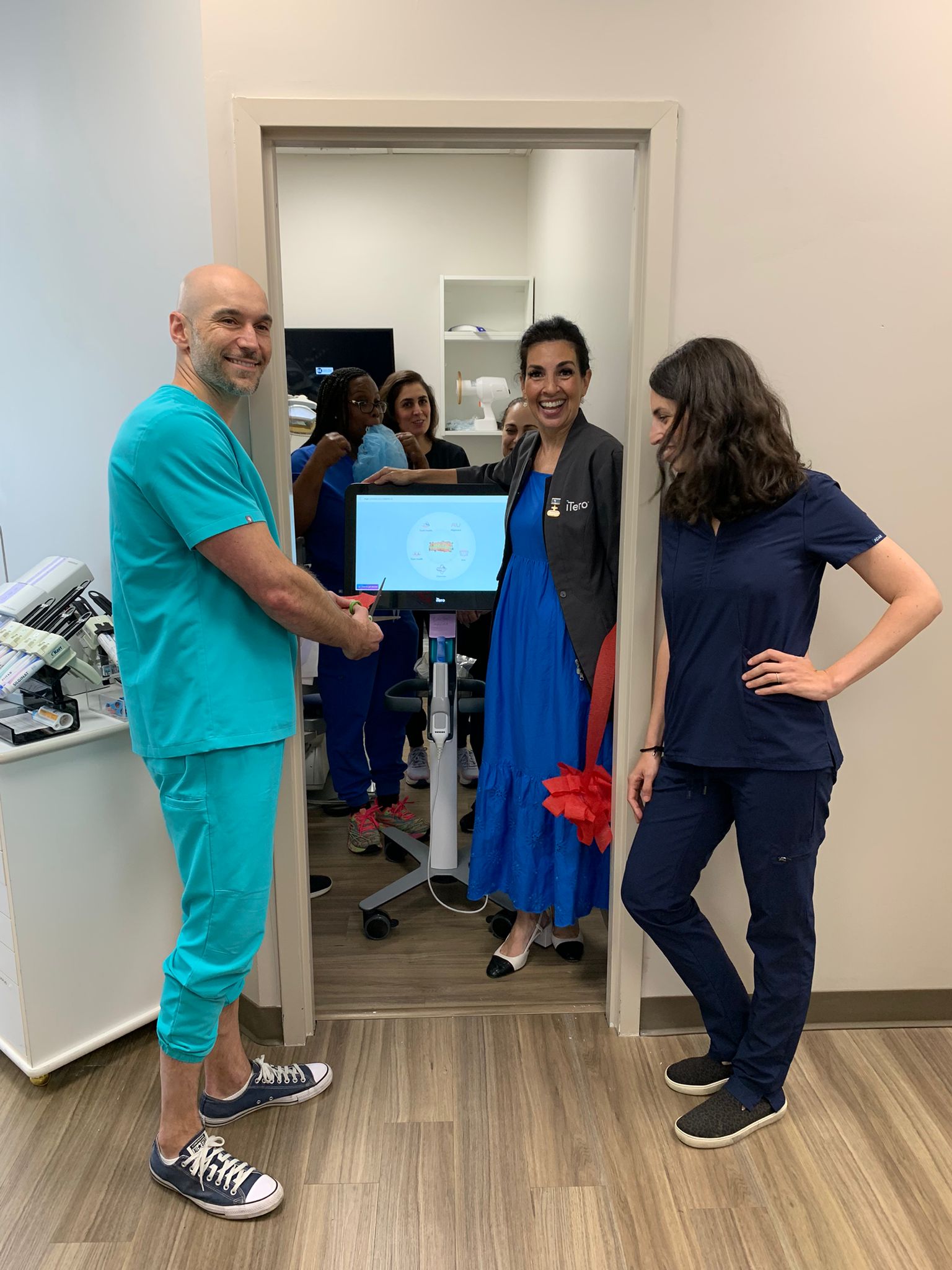 A group of dental professionals is gathered around an intraoral scanner in a dental office. A bald man in turquoise scrubs and Converse sneakers is smiling and pointing at the screen, while a woman in a blue dress with a black jacket, labeled “iTero,” stands beside him, also smiling. Another woman in navy blue scrubs is standing nearby, looking at them. In the background, several other team members are visible in the reflection of a mirror, watching the moment. The setting is a well-lit dental office with clean, professional surroundings. A red ribbon is tied to the scanner, suggesting a special occasion or equipment unveiling.