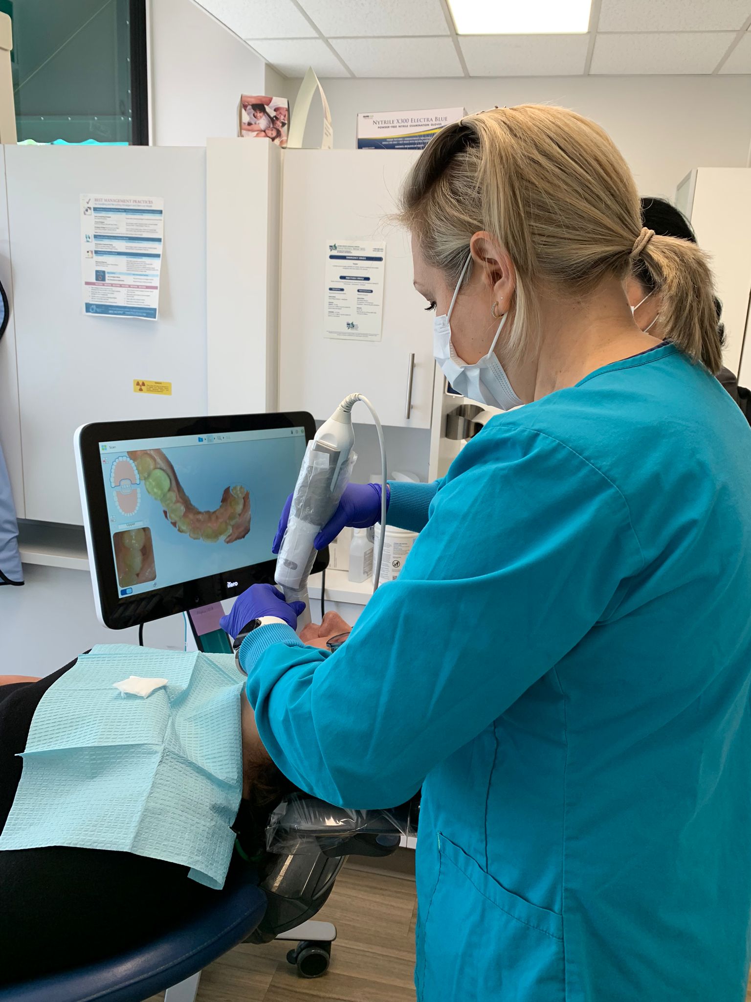 A dental assistant in a turquoise uniform and a face mask is performing a digital intraoral scan on a patient using a handheld scanner. The patient is lying back in a dental chair with a protective bib. The assistant is wearing purple gloves and is focused on capturing a 3D image of the patient’s teeth, which is displayed on a computer screen in front of her. The dental office environment is clean and professional, with white cabinets and dental equipment visible in the background.