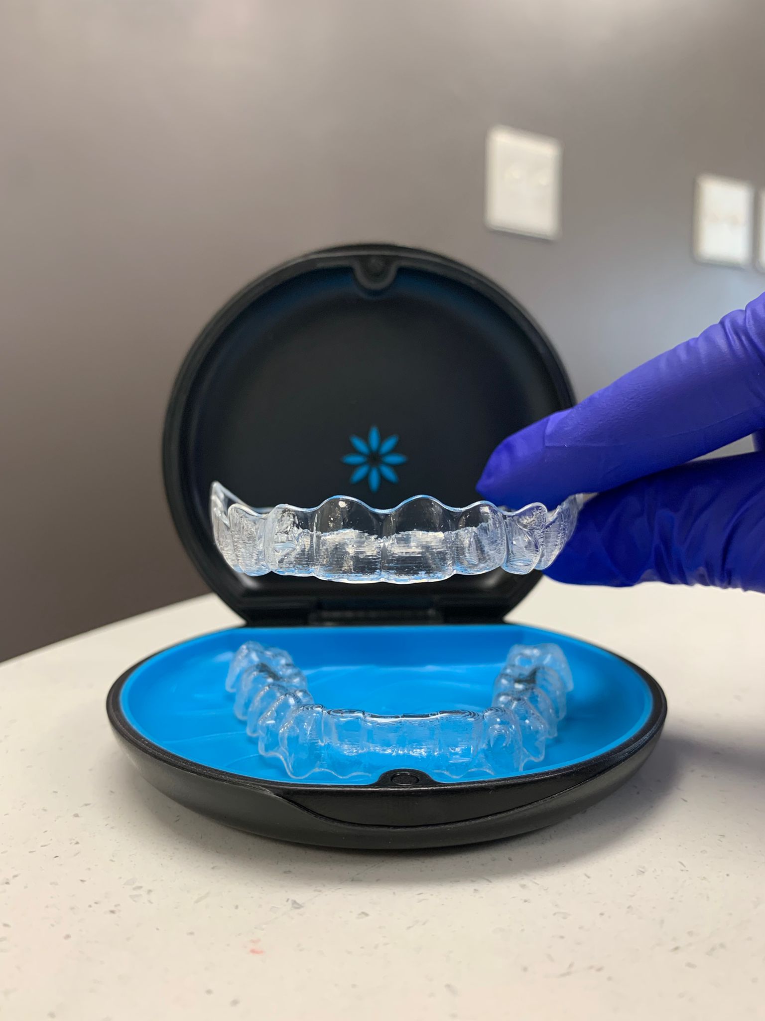 A close-up of a pair of clear aligners in a black and blue protective case. A gloved hand in blue latex holds the upper aligner above the case, showcasing its transparency and precise fit. The background features a neutral-colored wall with electrical outlets slightly blurred. The image highlights orthodontic treatment with clear aligners for teeth straightening and retention.