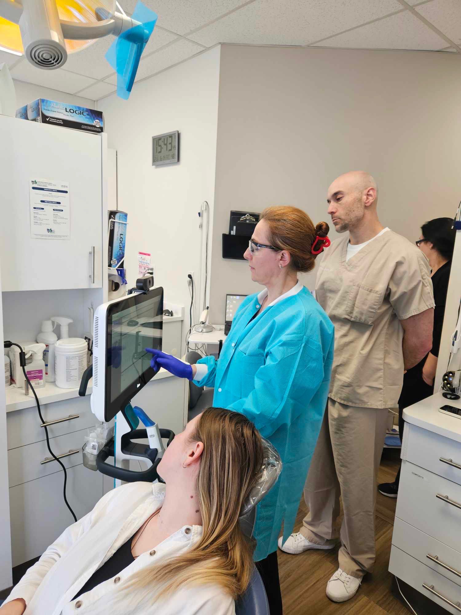 A dental professional in a blue protective gown and purple gloves is interacting with a touchscreen monitor, showing a patient seated in a dental chair some information. The patient, a woman with long blonde hair, is attentively looking at the screen. A male dental professional in beige scrubs stands behind them, observing the procedure. The dental clinic setting includes various dental tools, supplies, and equipment, creating a clean and professional environment.