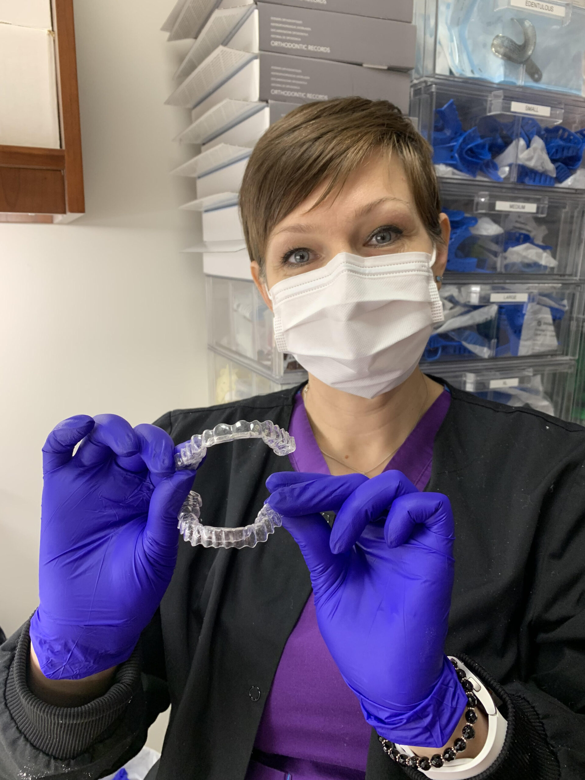 A dental professional wearing a black jacket, purple scrubs, a face mask, and blue gloves is holding up a set of clear retainers with both hands. She has short brown hair and is looking directly at the camera with a friendly expression. Behind her, shelves are stocked with orthodontic supplies, including labeled boxes and trays containing gloves and dental impressions. The setting is a clean and organized dental office, emphasizing professional orthodontic care.